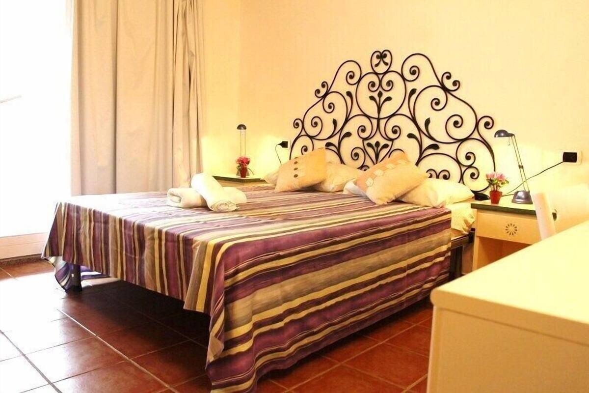 A bed with a striped bedspread, a decorative headboard, bedside lamps, and a nightstand with a vase of flowers.