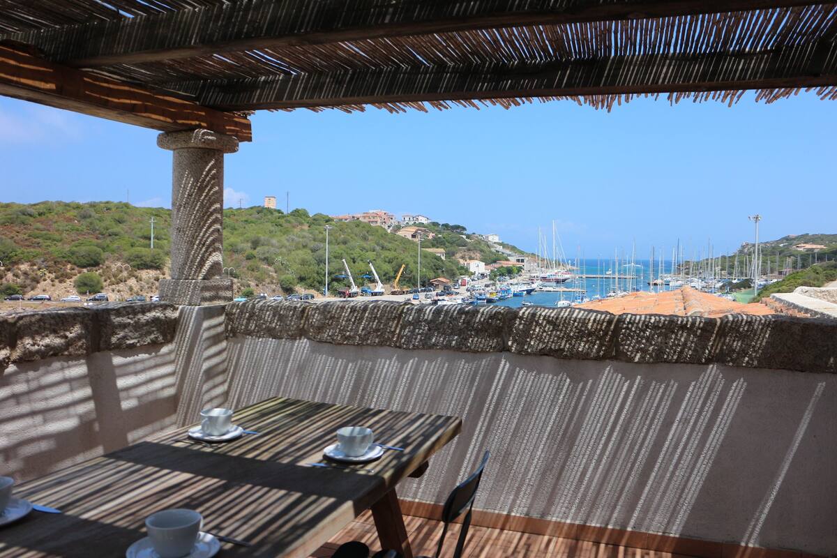 A terrace with a table set for dining, overlooking a marina with boats and a hilly landscape in the background.