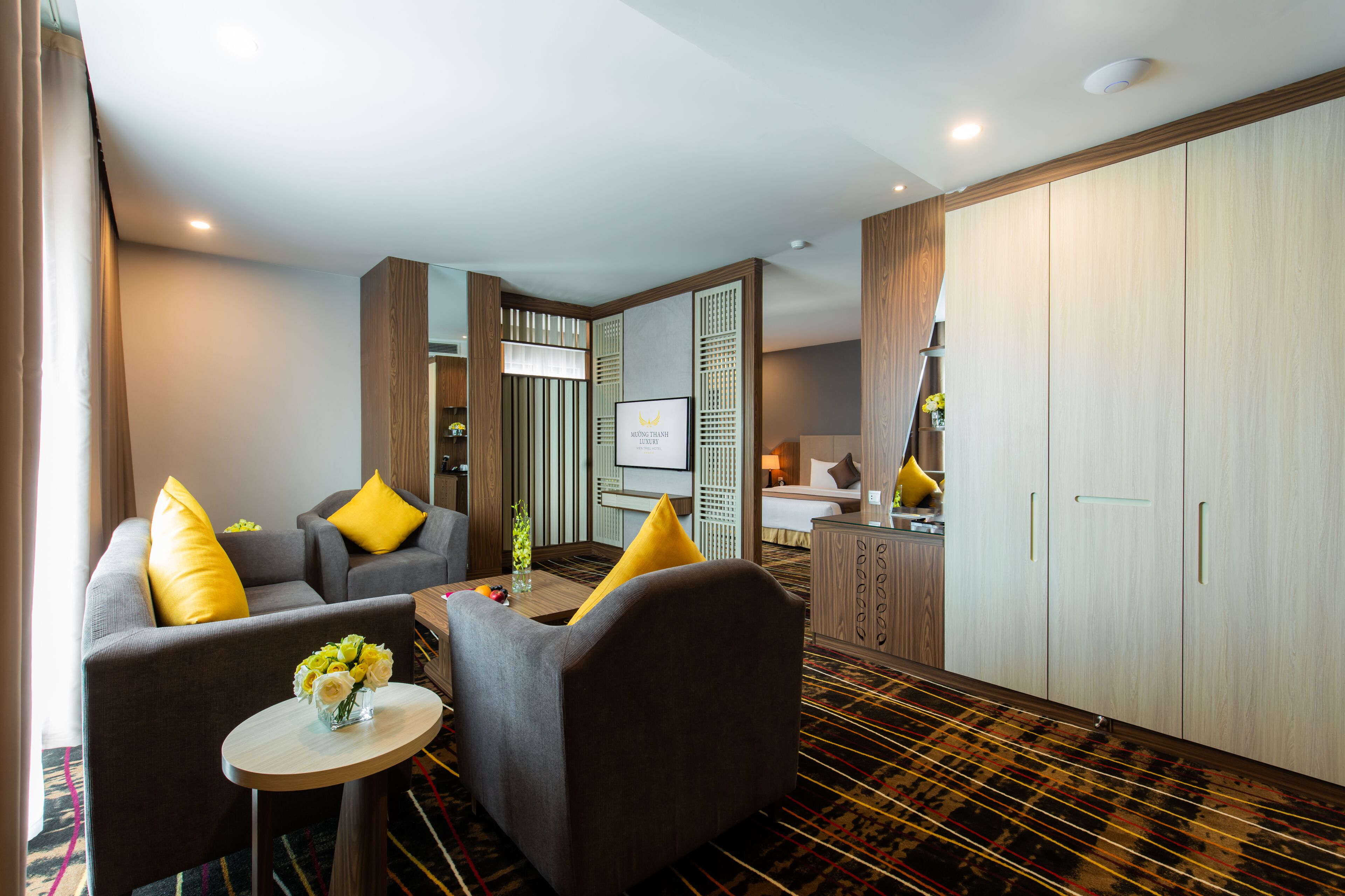 A hotel room with a sofa set, a small table with a vase of flowers, a bed, a wardrobe, and a television.