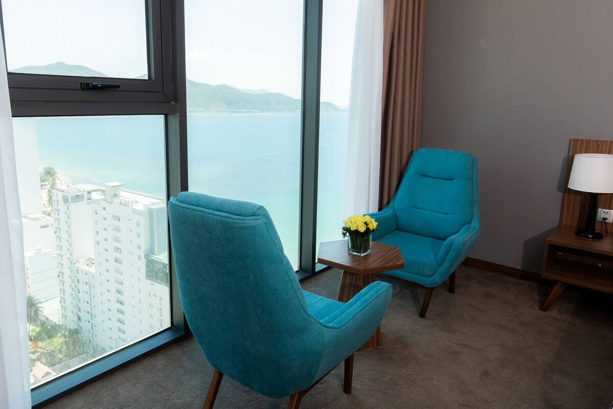 Une chambre d’hôtel dotée d’une grande fenêtre donnant sur la mer et les bâtiments, d’un fauteuil turquoise, d’une petite table sur laquelle est posé un vase rempli de fleurs jaunes, et d’une lampe de chevet.