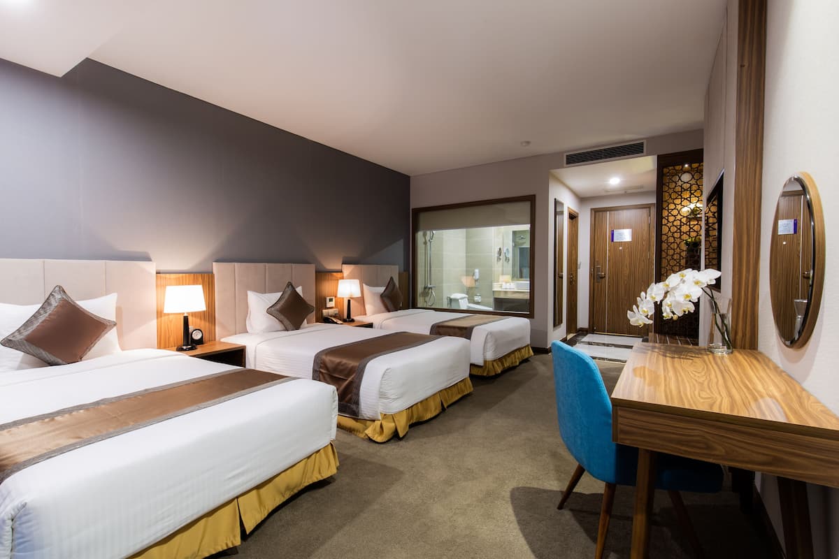 Une chambre d’hôtel avec trois lits, un bureau en bois, une chaise bleue et un miroir.