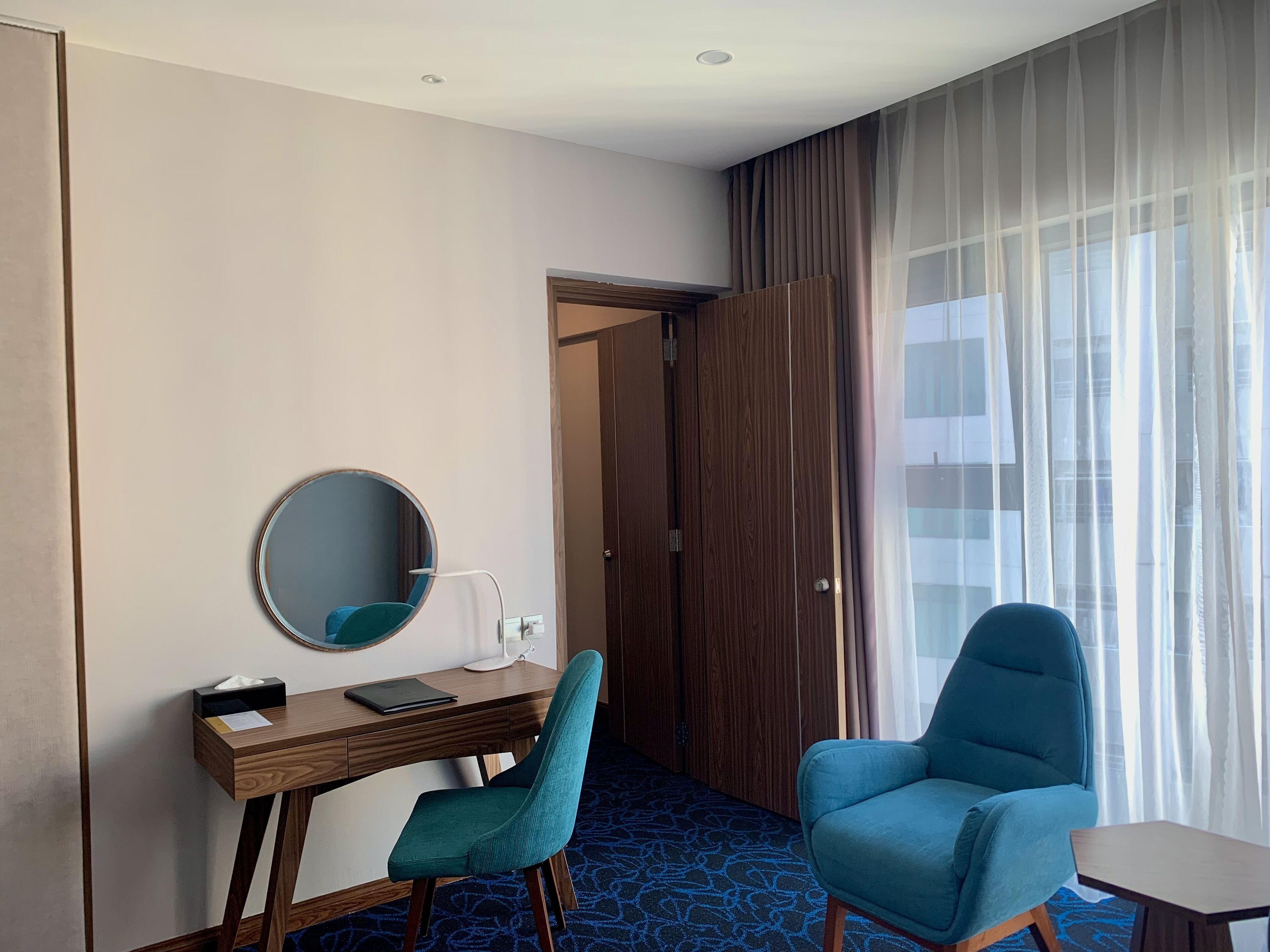 A hotel room with a wooden desk, a teal chair, a round mirror, and a large window with sheer curtains.