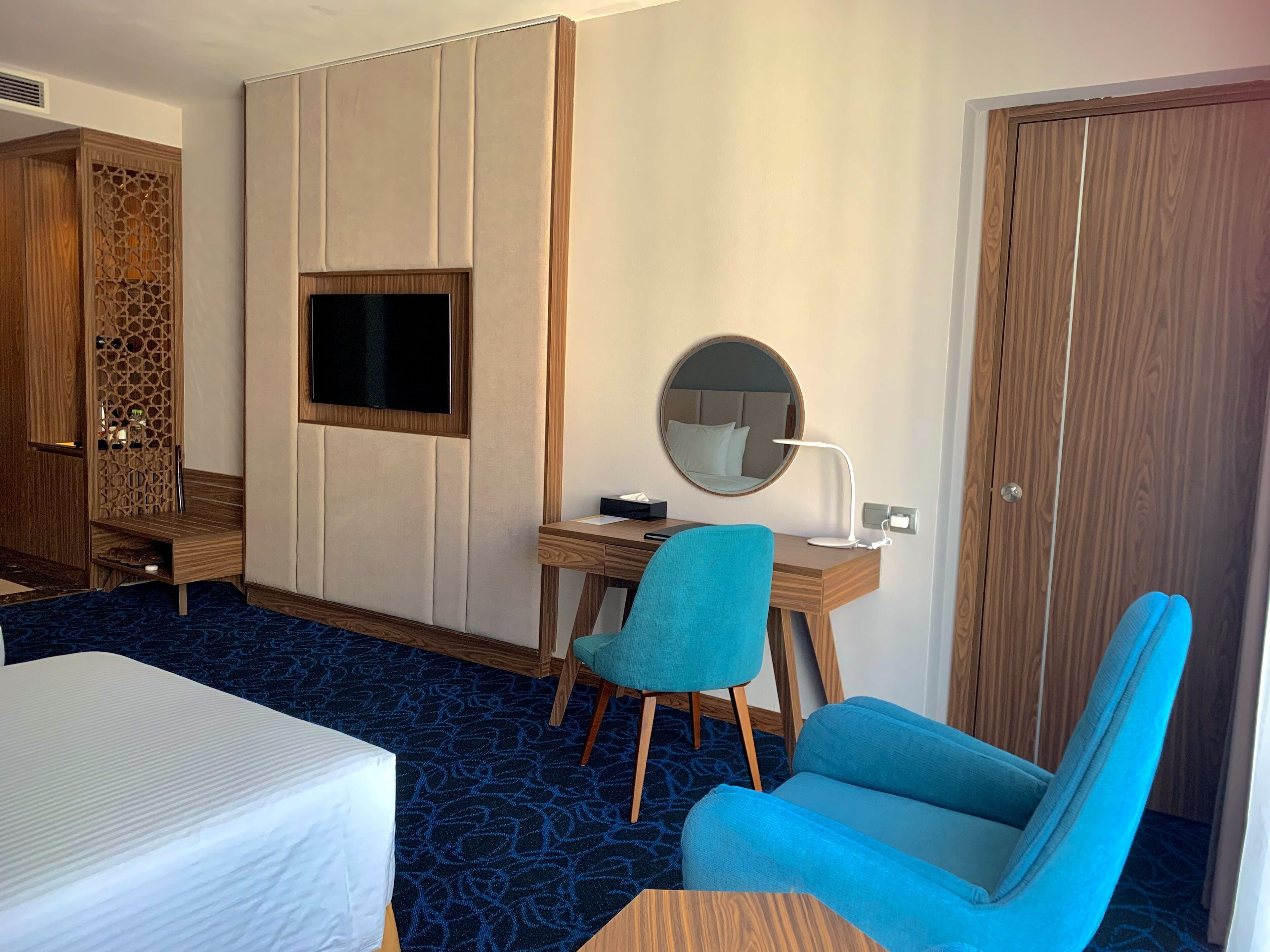 A hotel room with a bed, a desk with a chair, a TV, a mirror, and a wardrobe.