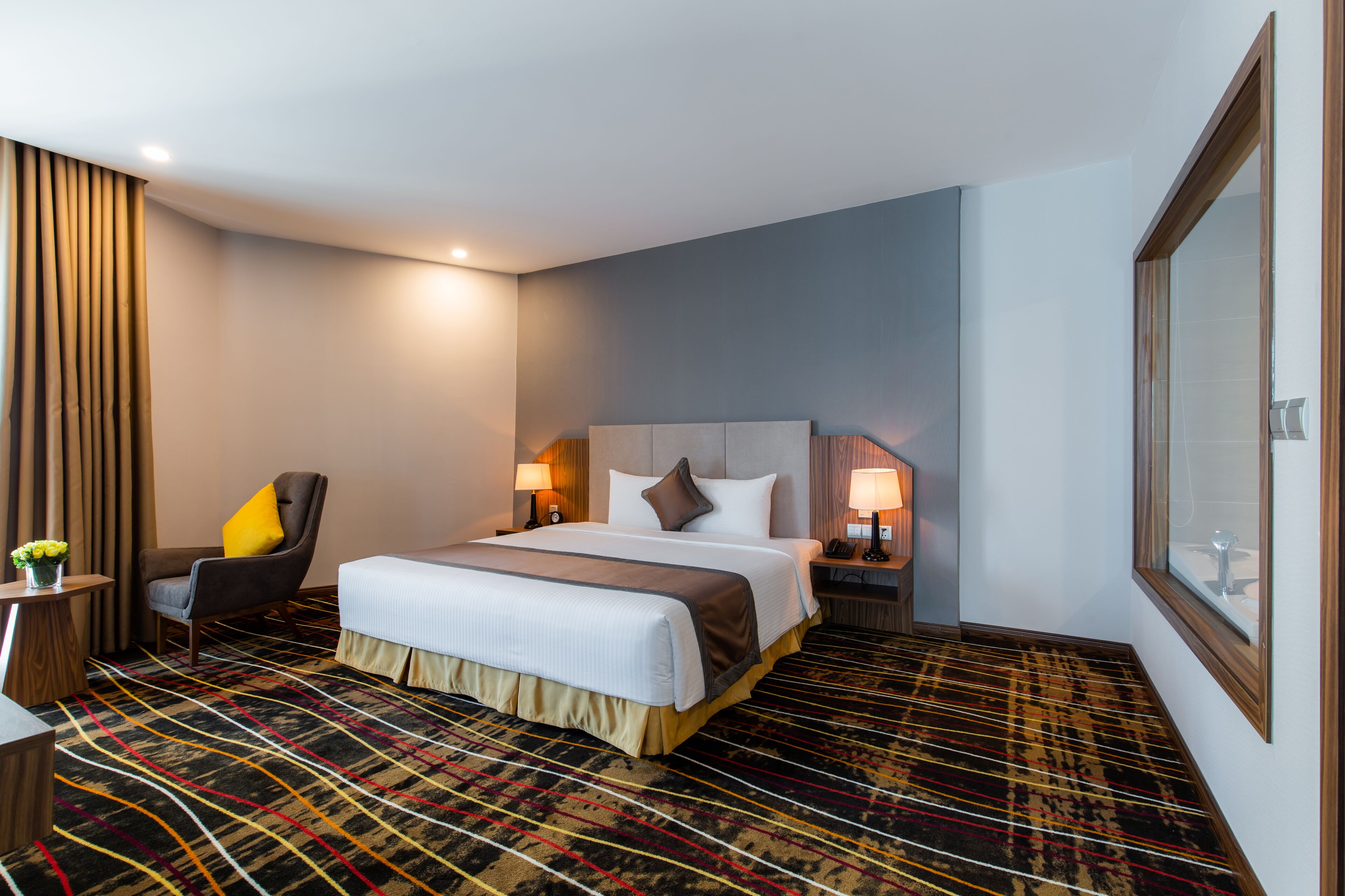 Hotellihuone, jossa on sänky, tuoli, yöpöytä, lamppu ja suuri peili.