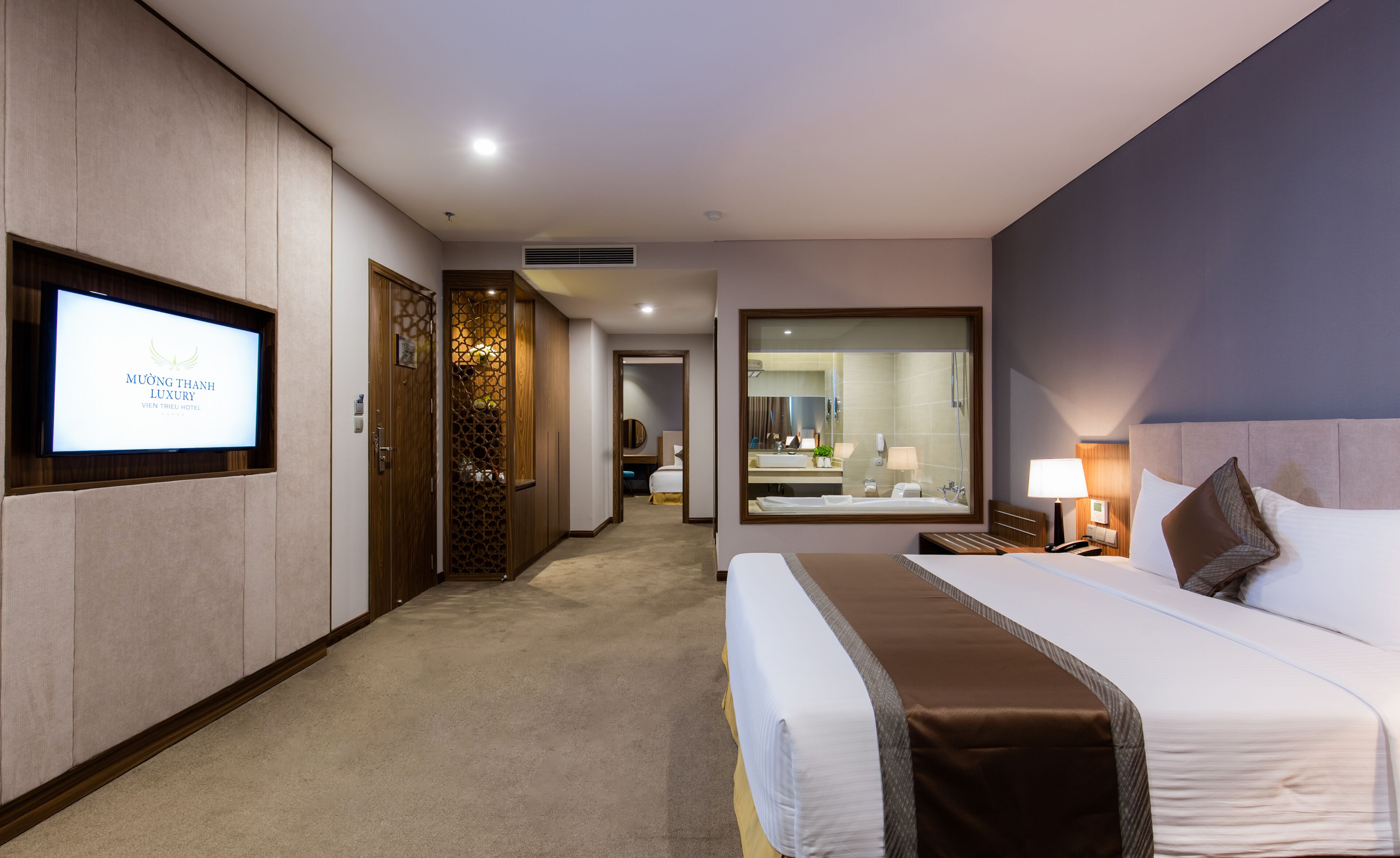 A hotel room with a large bed, a television mounted on the wall, a bathroom with a mirror, and a hallway leading to another room.