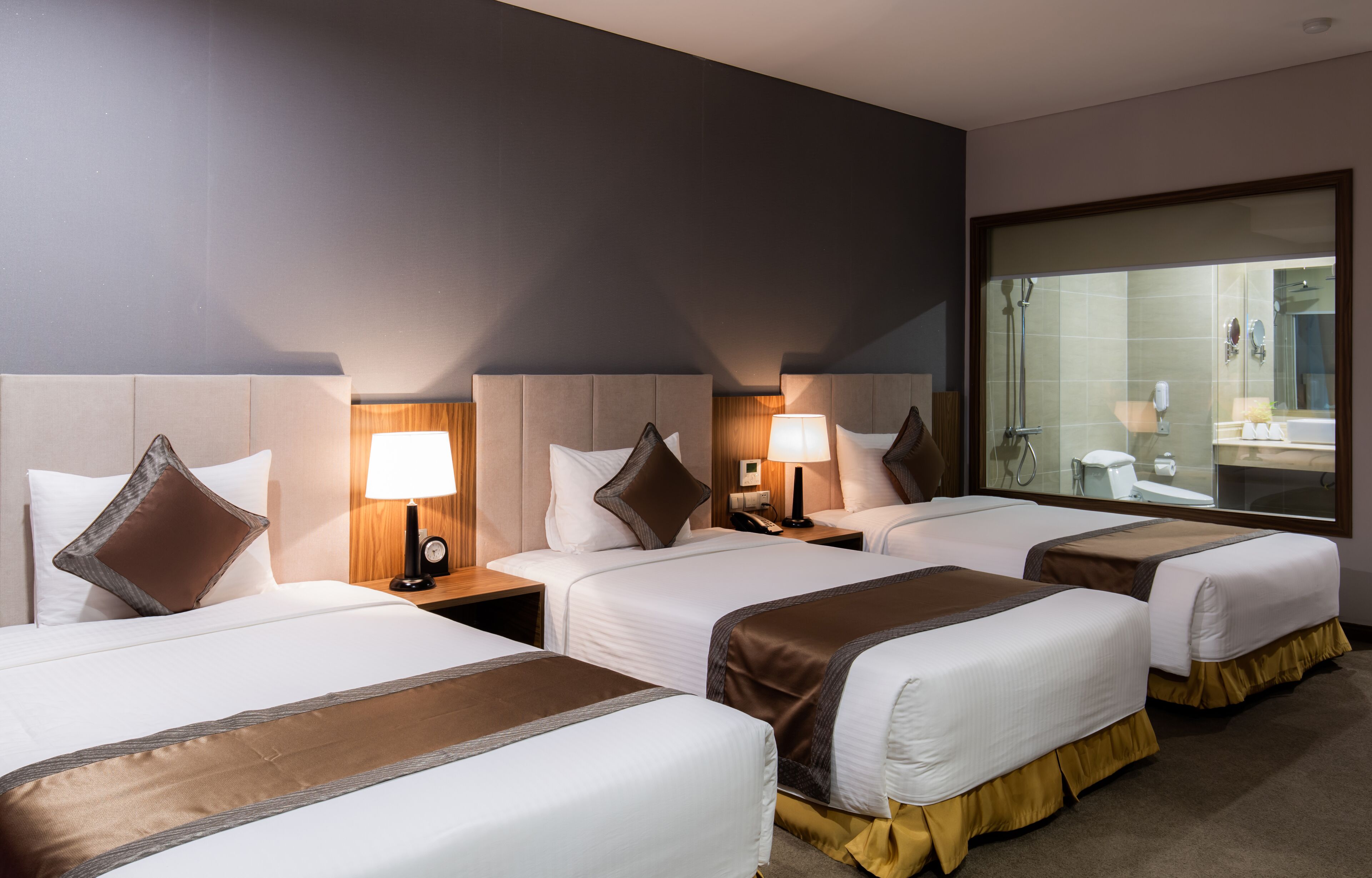 A hotel room with three beds, each with a pillow and a bedside table with a lamp. There is a large mirror on one wall and a bathroom visible through a glass door.