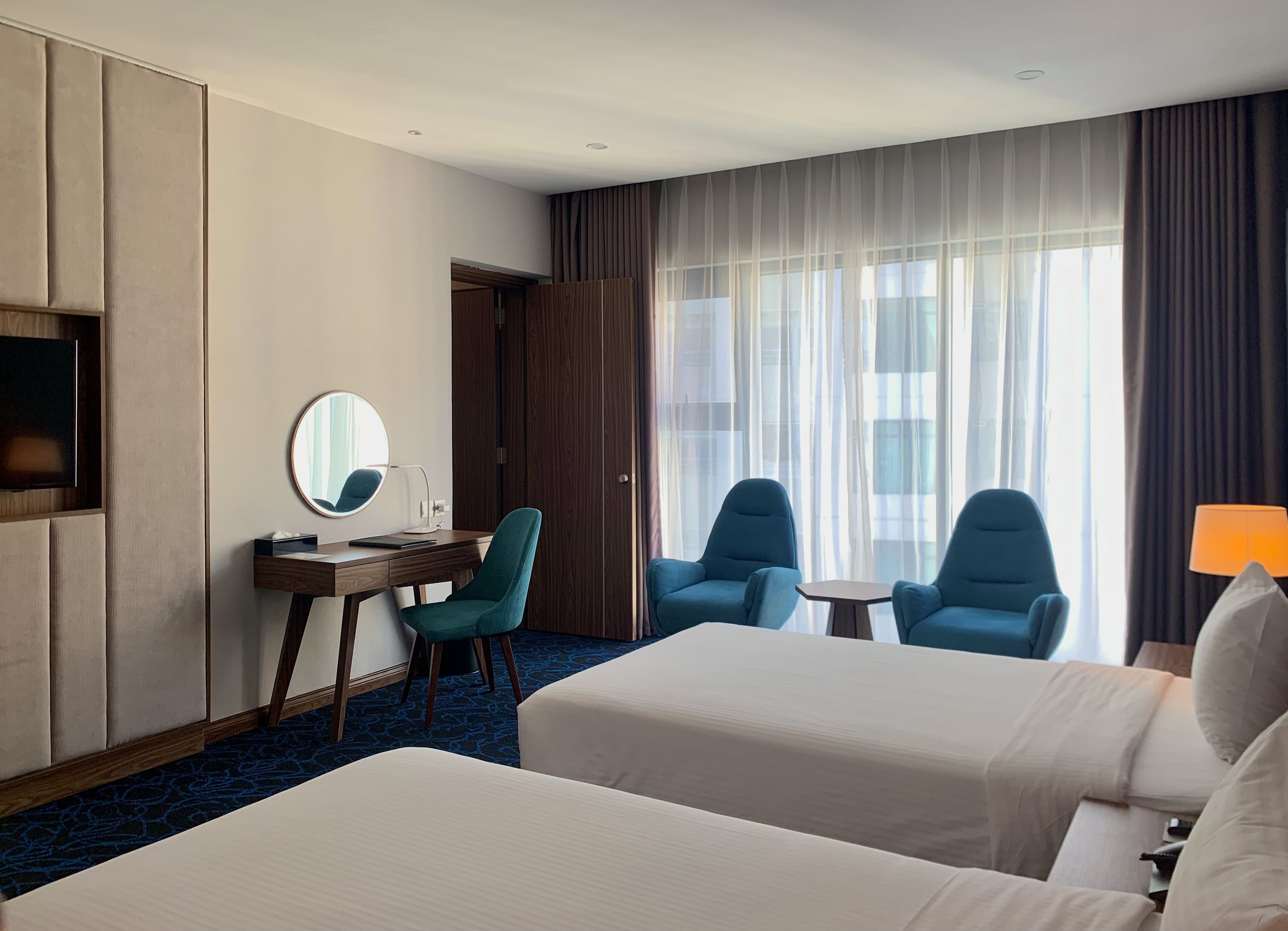 A hotel room with two beds, a desk with a chair, a TV, and a large window with curtains.