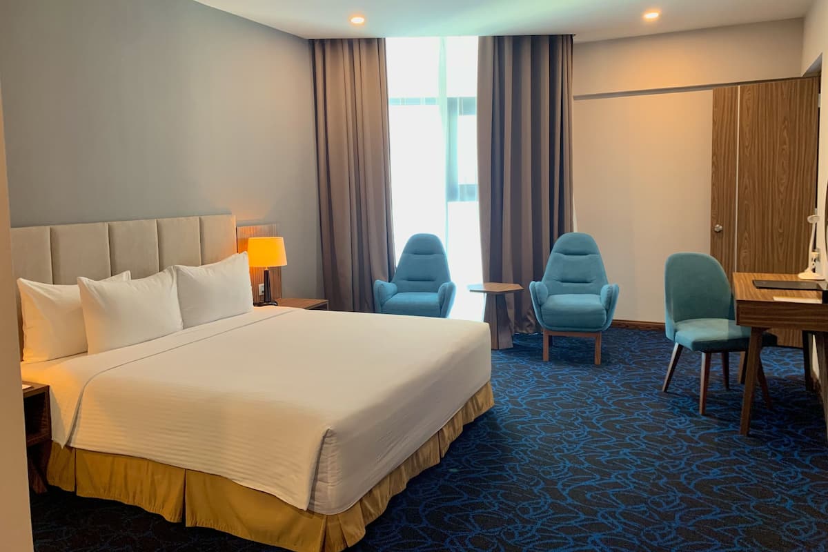 Une chambre d’hôtel avec un grand lit, deux fauteuils, une petite table et un bureau avec une lampe.