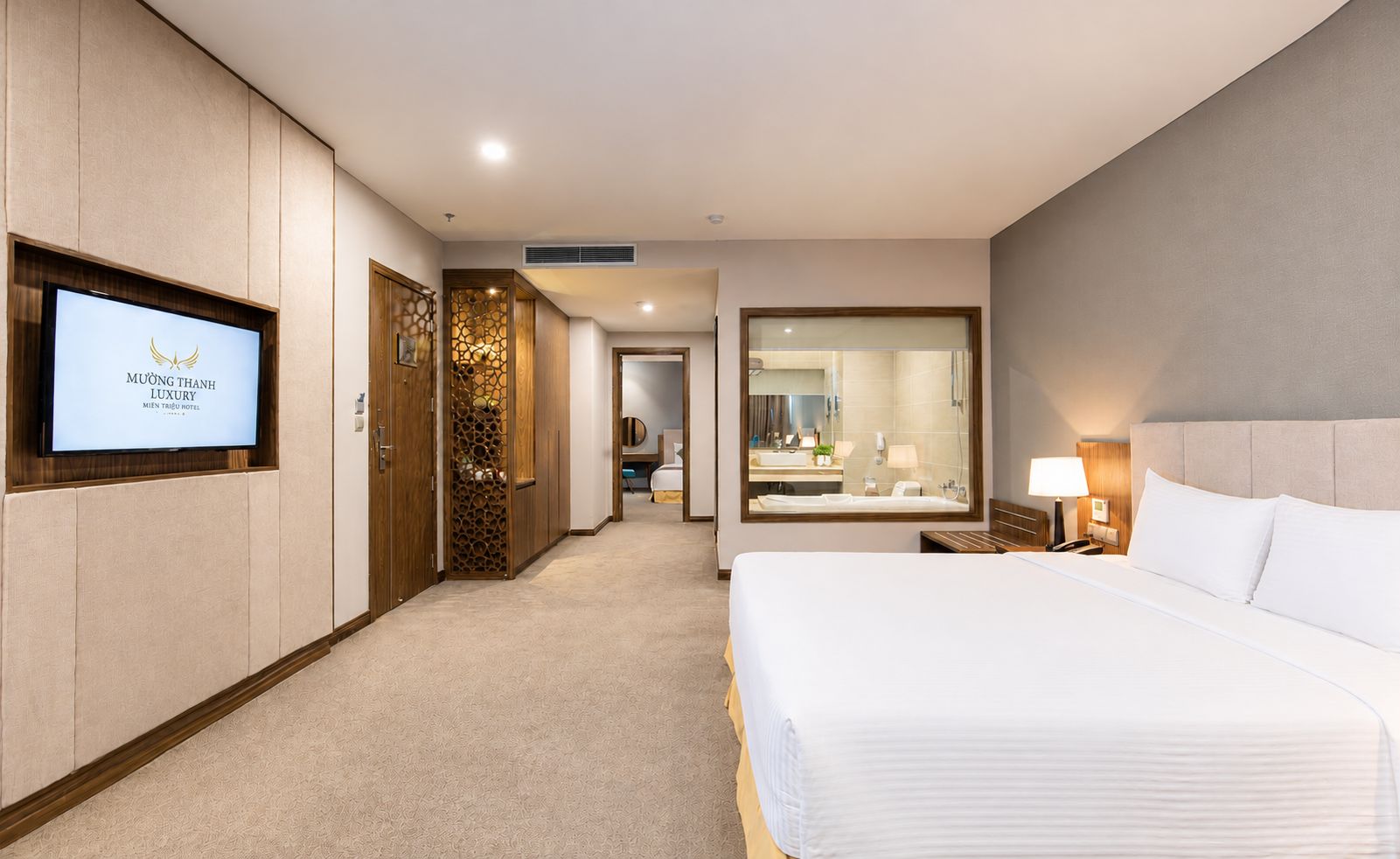 Habitación de hotel con cama, televisor montado en la pared, baño con espejo y un pasillo que conduce a otra habitación.