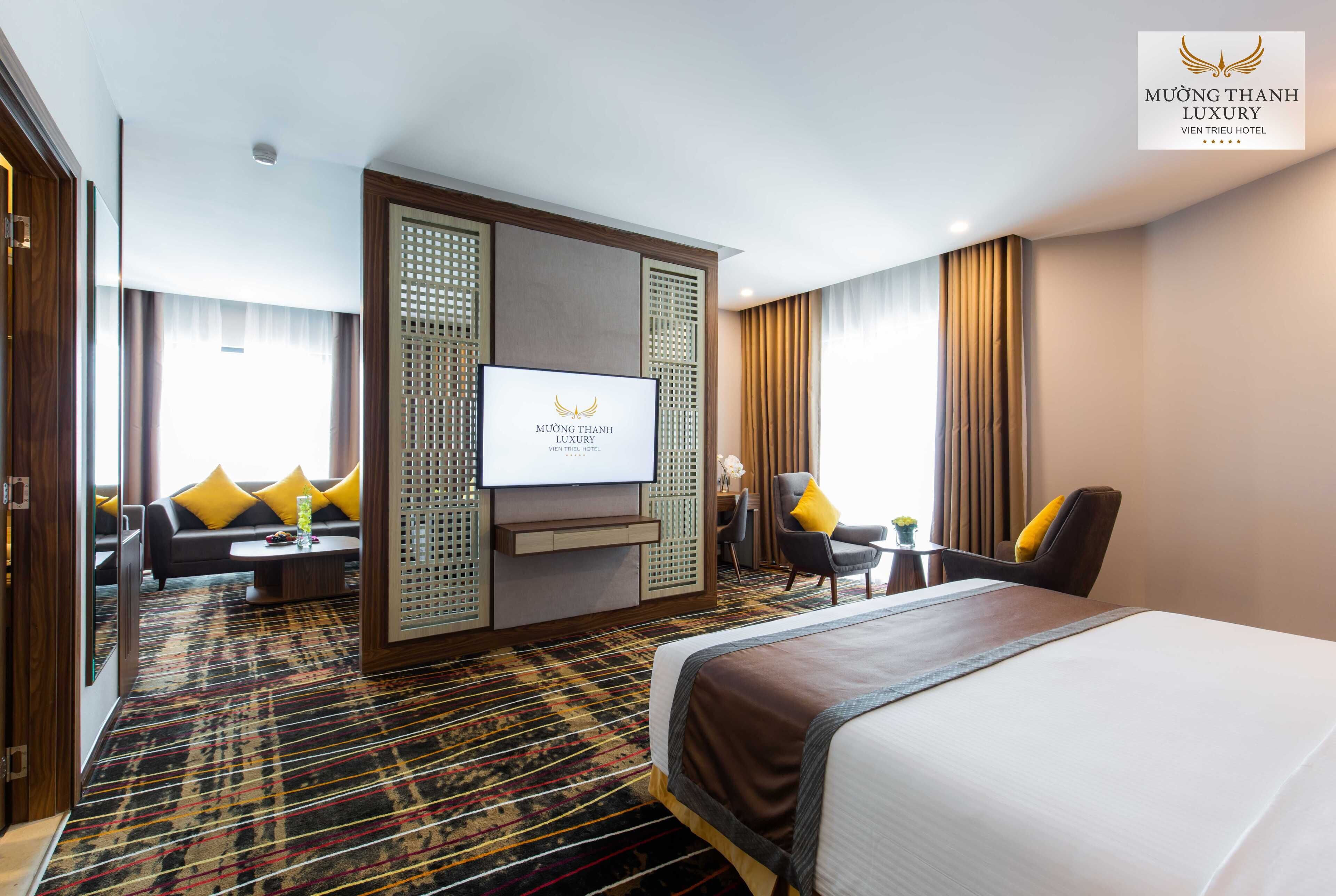 Una habitación de hotel con una cama grande, un televisor, un sofá con cojines amarillos, un escritorio con una silla y una alfombra estampada.