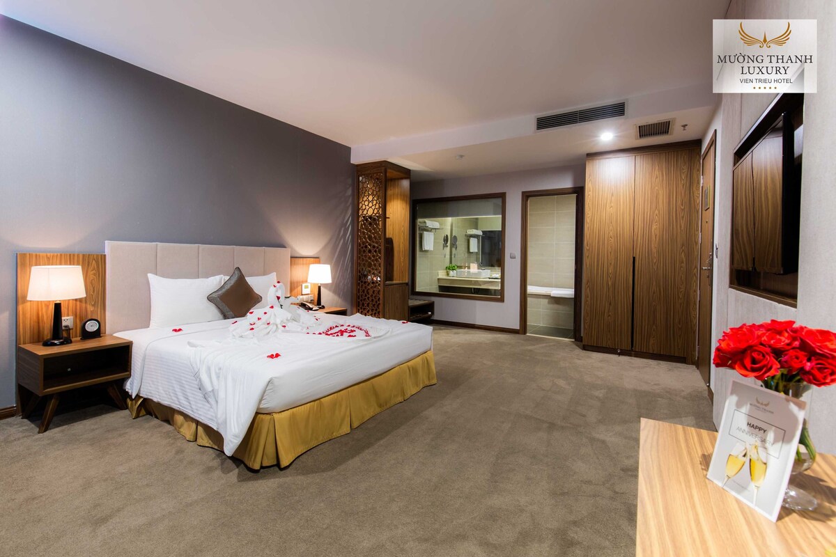 Une chambre d’hôtel avec un grand lit, des lampes de chevet, une armoire en bois, un miroir et une petite table sur laquelle est posé un vase de fleurs.