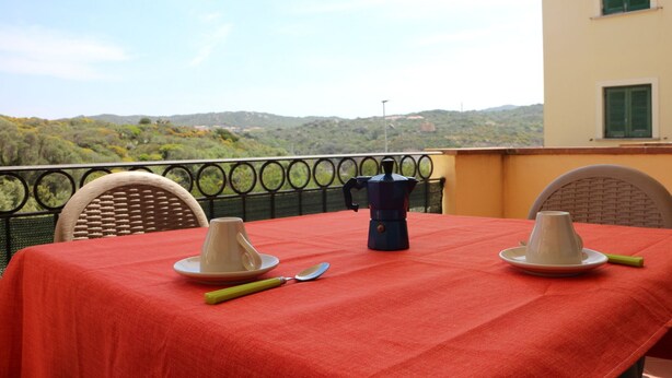 A table set with a coffee pot, cups, and a spoon on a red tablecloth, overlooking a scenic landscape. - Residence Lampara (Santa Teresa di Gallura)