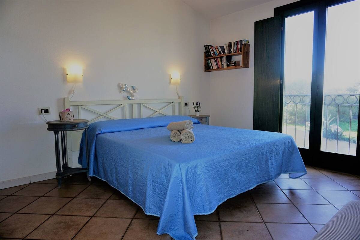 A bedroom with a bed, two bedside tables, a nightstand, a bookshelf, and a window with a view.