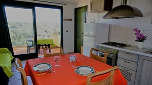 A dining area with a table set for two, a kitchen with a gas stove, and a view of the outdoors through a sliding door. - Residence Lampara (Santa Teresa di Gallura)