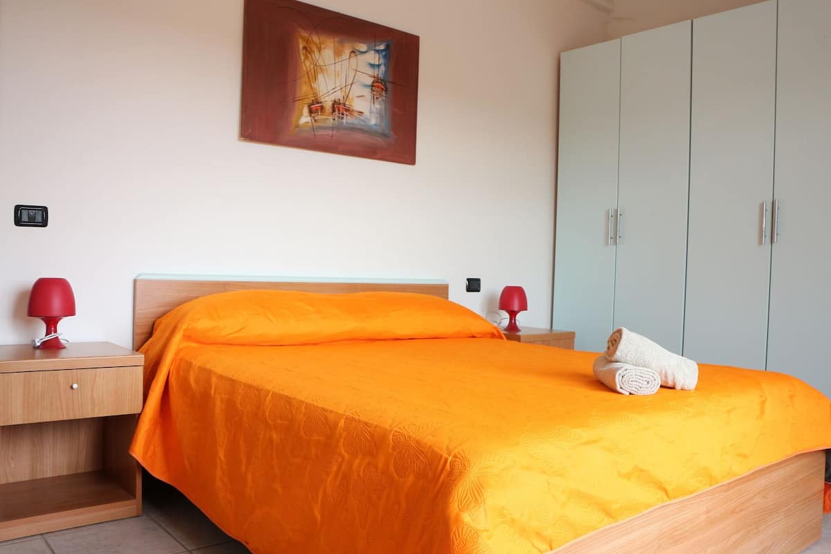 A neatly made bed with an orange bedspread, two bedside tables with red lamps, a painting on the wall, and a wardrobe.