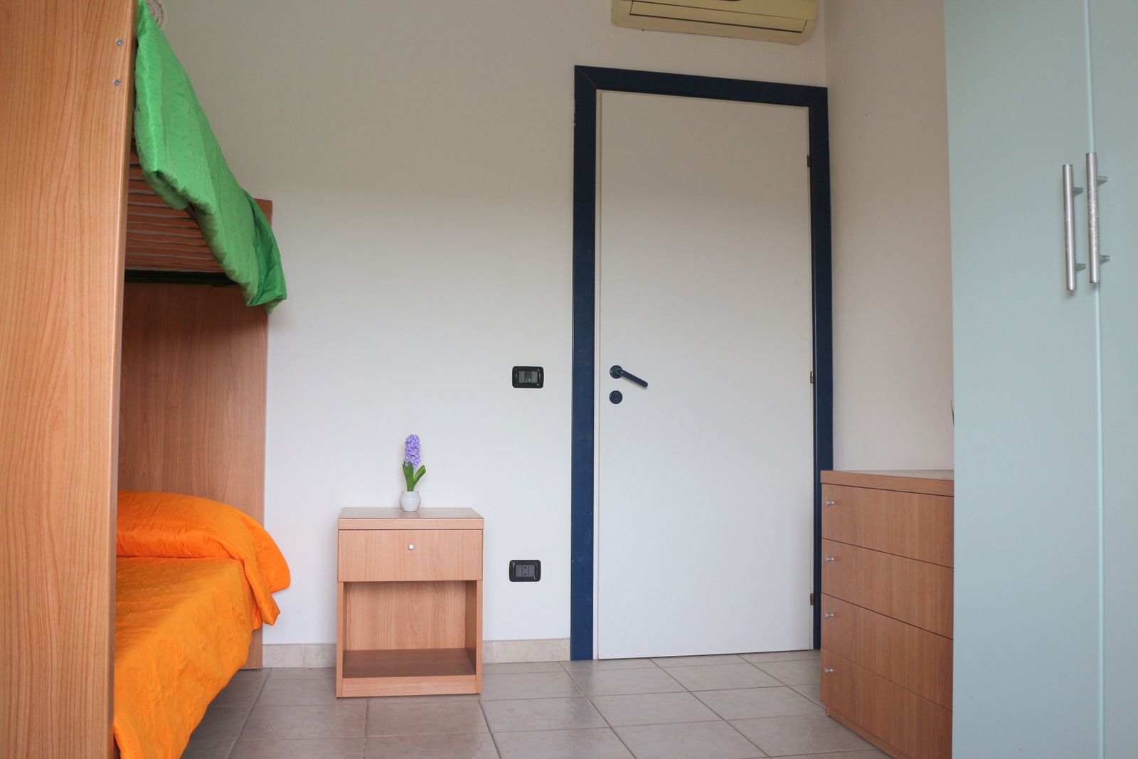 A room with a bunk bed, a small wooden cabinet, a door, and a wall-mounted air conditioner.