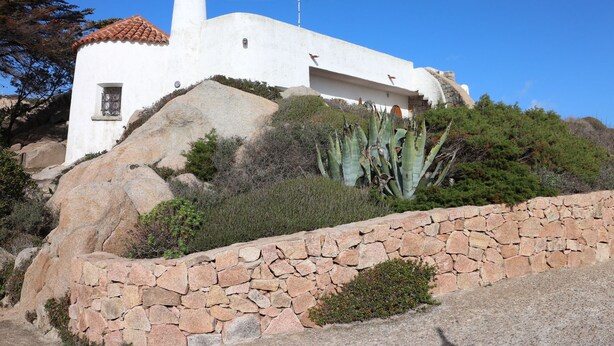 Un edificio blanco con techo de tejas rojas, ubicado en una colina con un muro de piedra en primer plano.