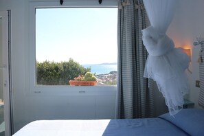 A bedroom with a large window, a bed, a nightstand, a potted plant, and a view of the sea and greenery. - Villette Valle dell'Erica (Santa Teresa di Gallura)