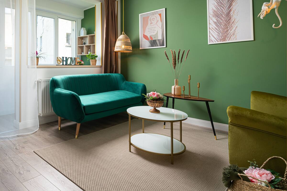 A living room with a teal sofa, a round coffee table, and a green armchair. There are two wall-mounted artworks and a small side table with a vase of dried plants.