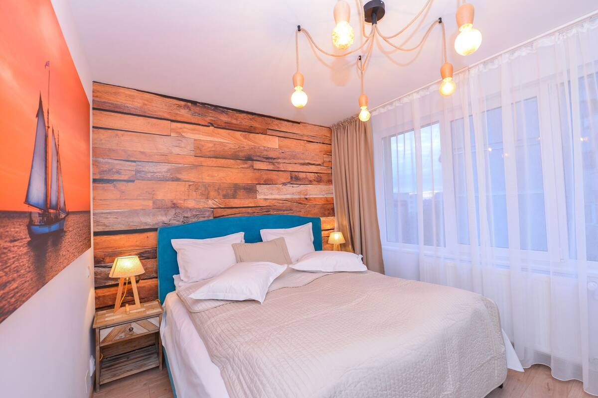 A bedroom with a wooden headboard, a bed with white bedding, a nightstand with a lamp, and sheer curtains on the window.