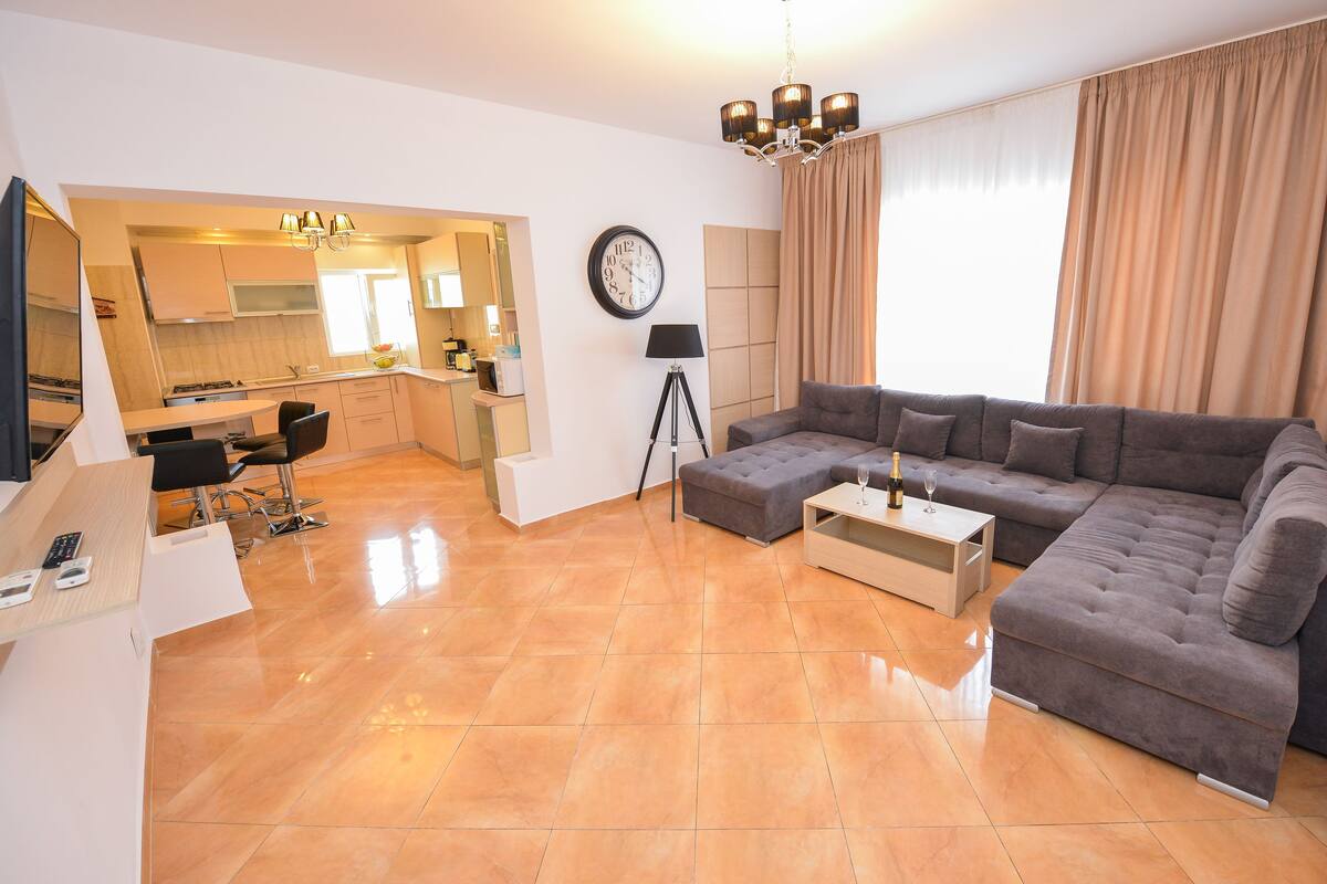 A modern living room with a sectional sofa, a dining area with a kitchen in the background, and a large window with curtains.