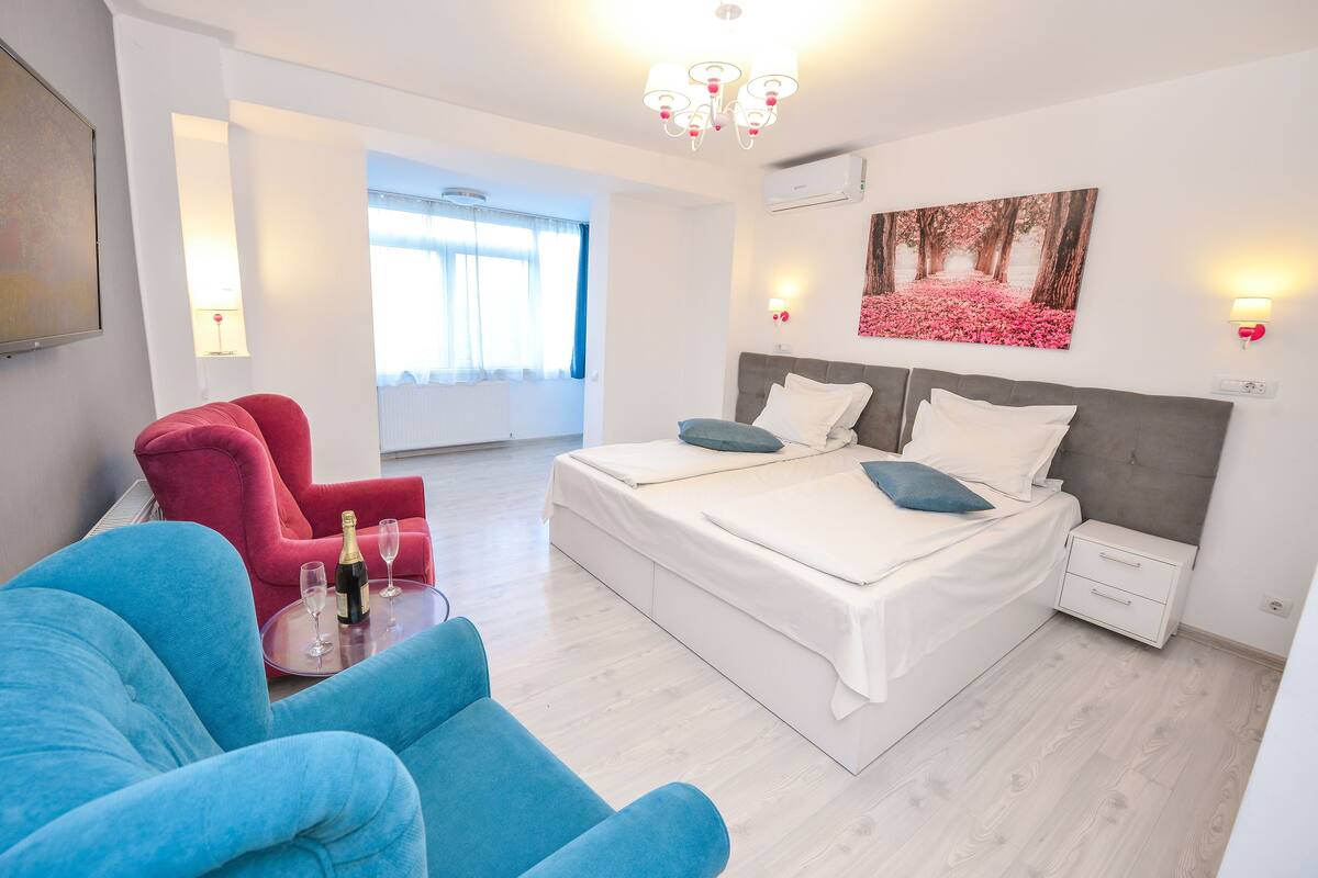 A modern hotel room with a large bed, a red armchair, a blue sofa, and a small table with a bottle and glass.