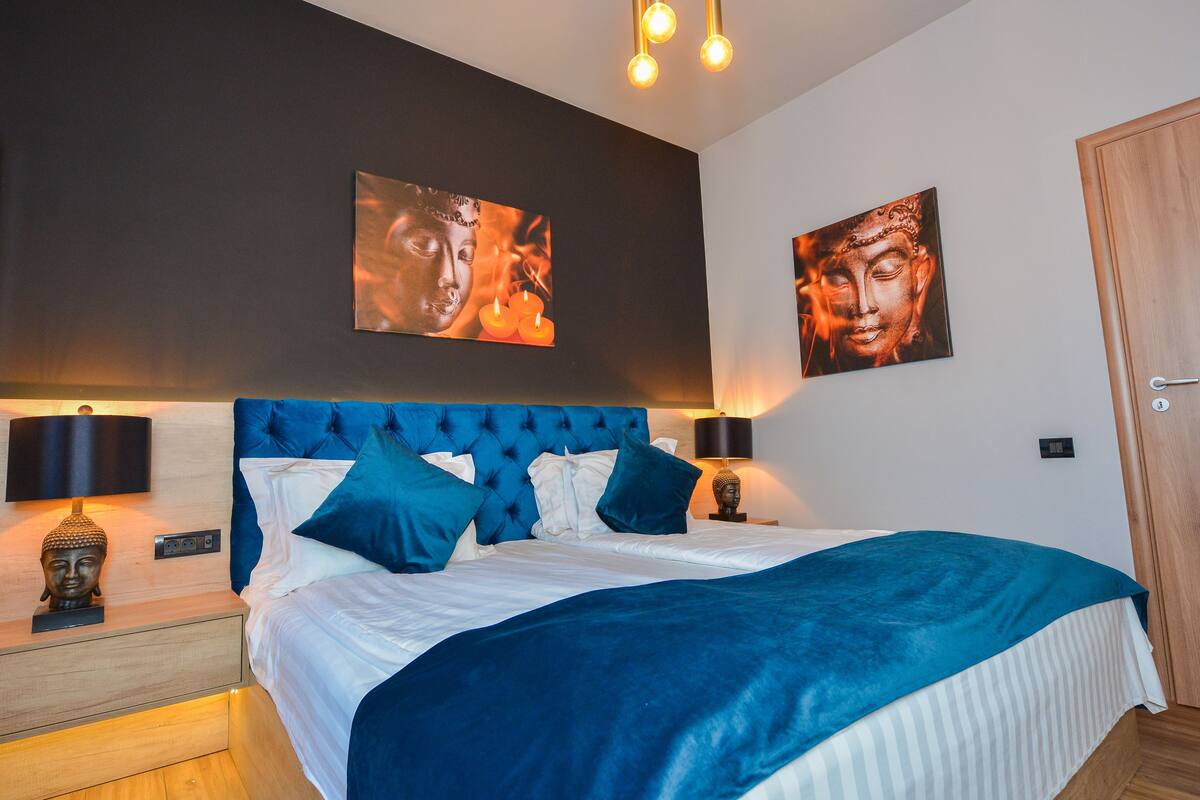 A bedroom with a large bed, two wall-mounted lamps, and two paintings.