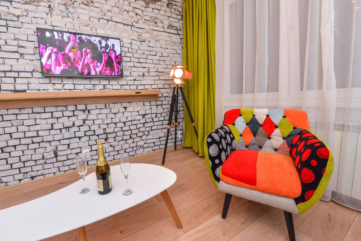 A modern living room with a brick wall, a mounted TV, a table with a bottle and glasses, a colorful armchair, and a floor lamp.
