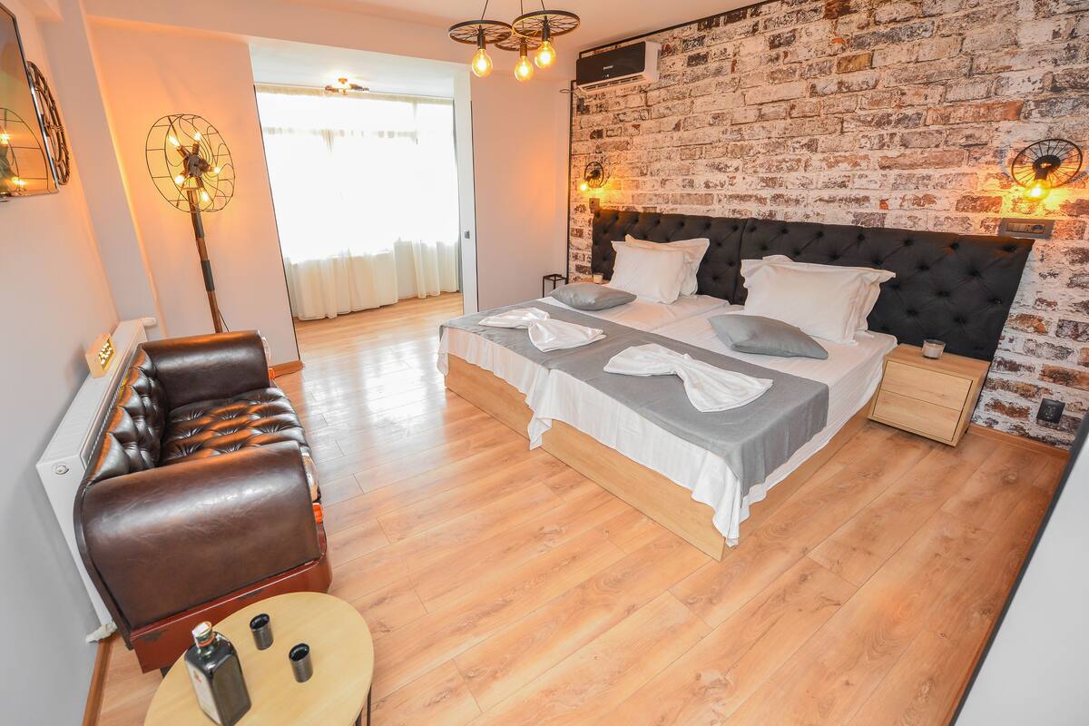 A modern bedroom with a brick-patterned wall, a tufted headboard, a leather sofa, and a wooden floor.