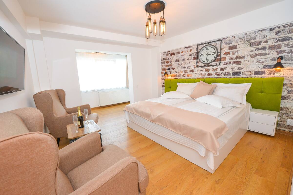A modern bedroom with a brick-patterned wall, a green upholstered headboard, a large bed, a flat-screen TV, and a seating area with two chairs.