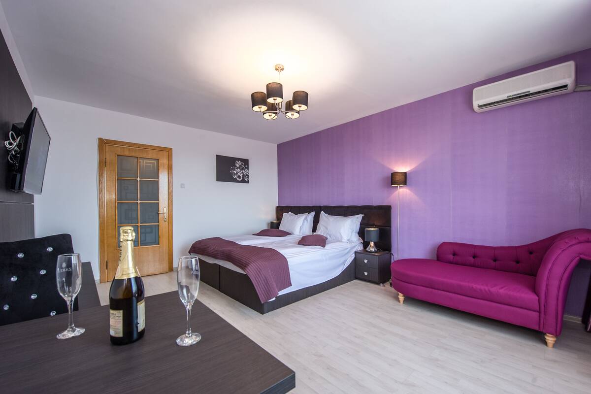 A modern bedroom with a bed, bedside tables, a purple sofa, a wooden door, and a table with glasses and a bottle.