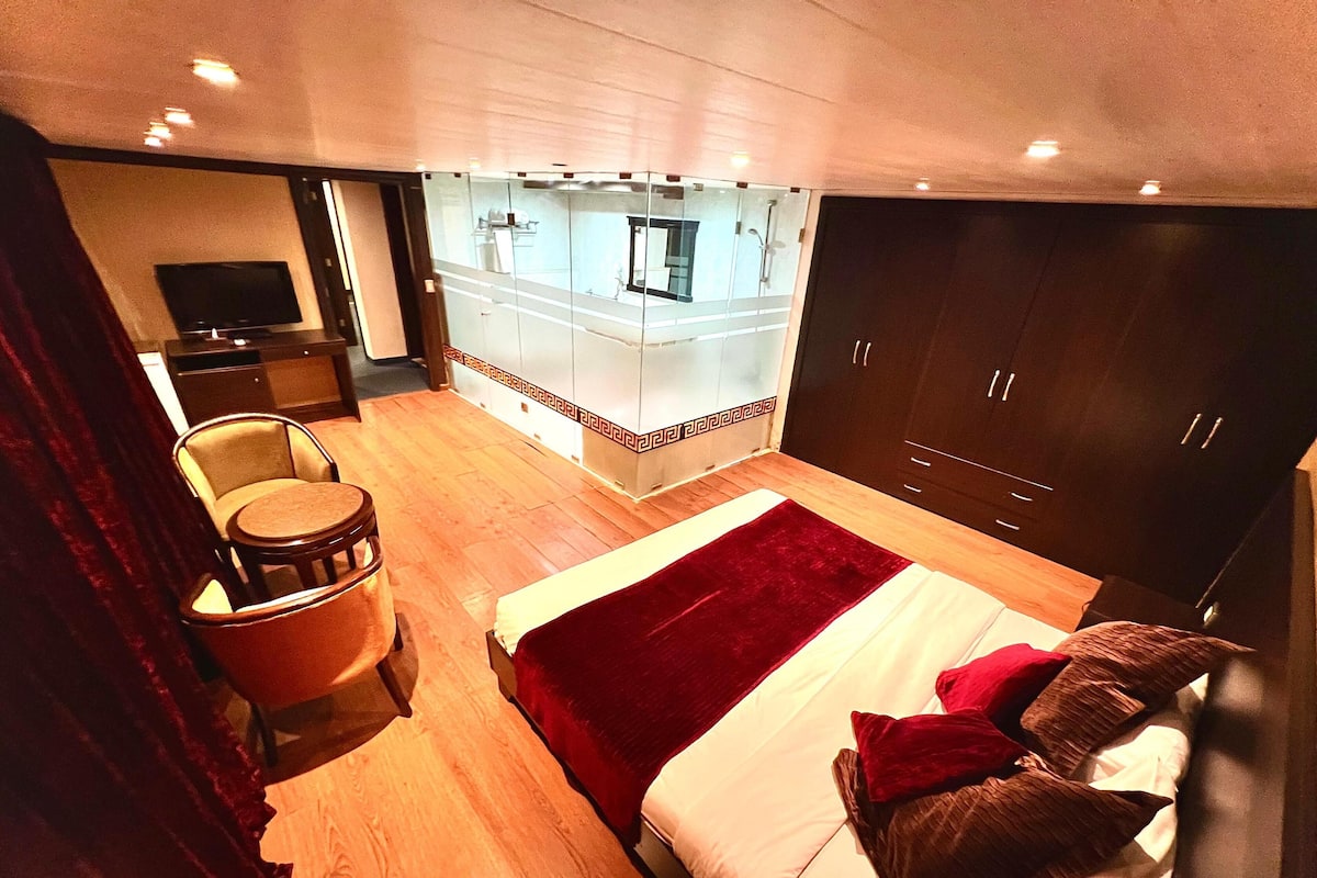 A modern hotel room with a large bed, a seating area with a chair and a small table, a television, and a bathroom visible through a glass partition.
