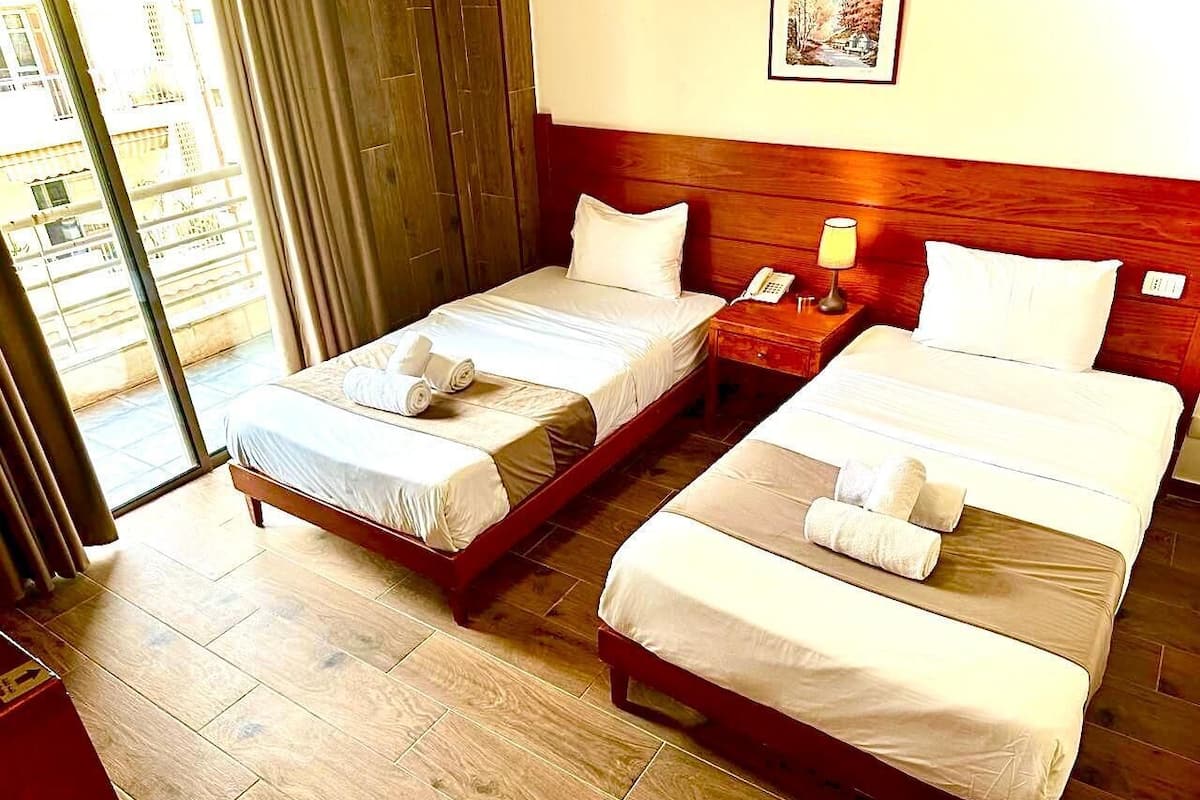 A hotel room with two beds, wooden flooring, a wooden headboard, a bedside table with a lamp, and a telephone.