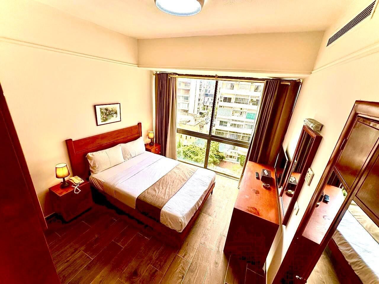A hotel room with a bed, bedside table, lamp, and a view of the city through large windows.