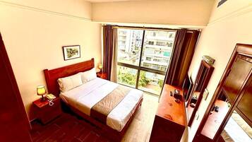 A hotel room with a bed, bedside table, lamp, and a view of the city through large windows.
