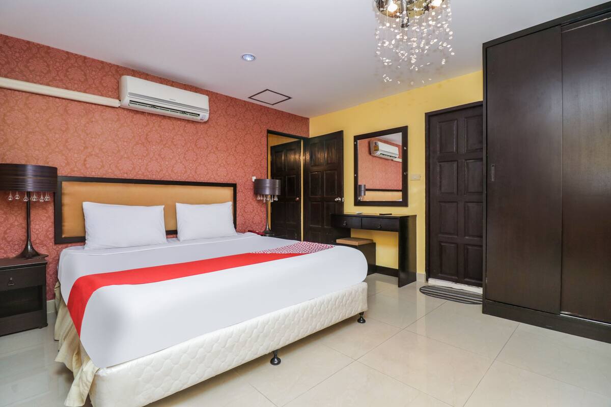 A hotel room with a large bed, a bedside table with a lamp, an air conditioning unit, a wardrobe, and a mirror.