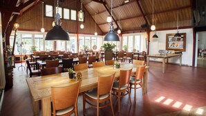 A spacious dining area with wooden tables and chairs, large hanging pendant lights, and a high ceiling with visible beams. - Buitengoed Fredeshiem (De Bult)