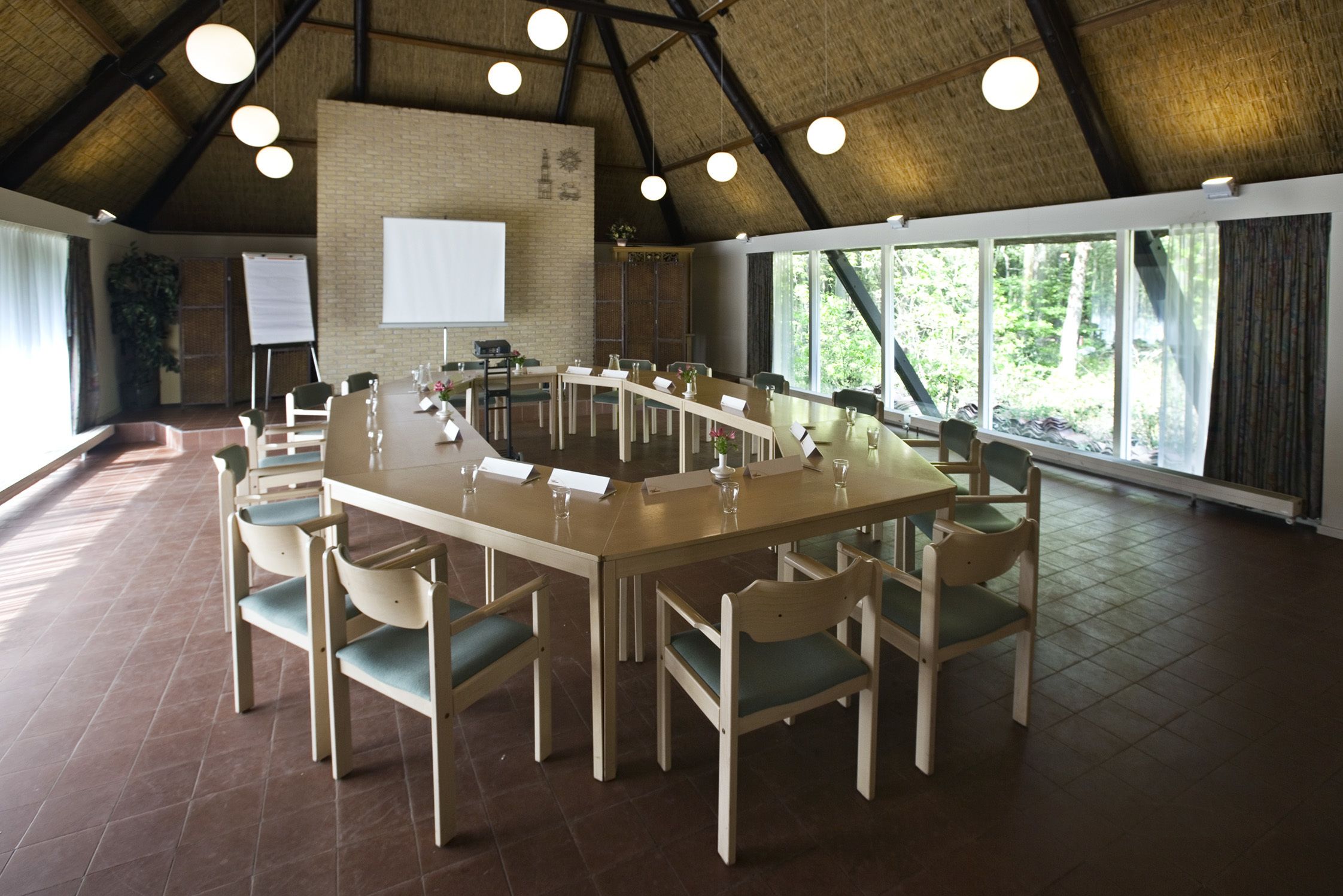A conference room with a long table, chairs, a projector screen, and a large window with a view of trees.