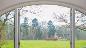 A view through a glass door leading to a paved area with a grassy field and trees in the background. - Buitengoed Fredeshiem (De Bult)