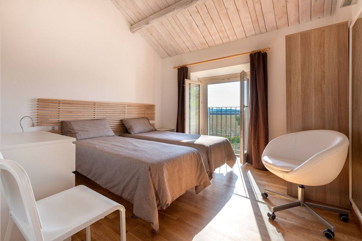 A bedroom with two beds, a desk, a chair, and a view of the outdoors.