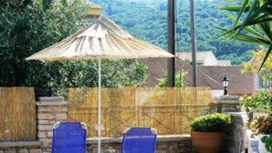 A patio with two lounge chairs and a sun umbrella, surrounded by a stone wall and a wooden fence. - Emily's Apartments (Corfu)