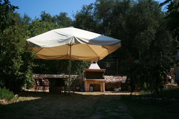 An outdoor patio with a stone barbecue grill and a table under a large umbrella. - Emily's Apartments (Corfu)