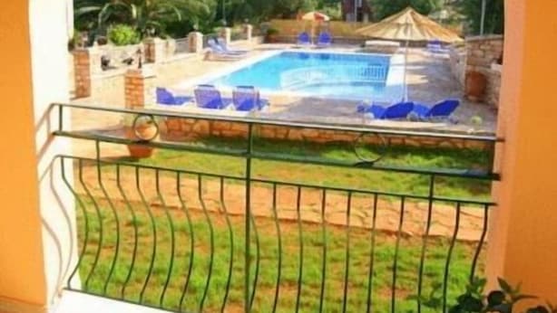 A balcony with a view of a swimming pool, surrounded by greenery and a distant mountain range. - Emily's Apartments (Corfu)