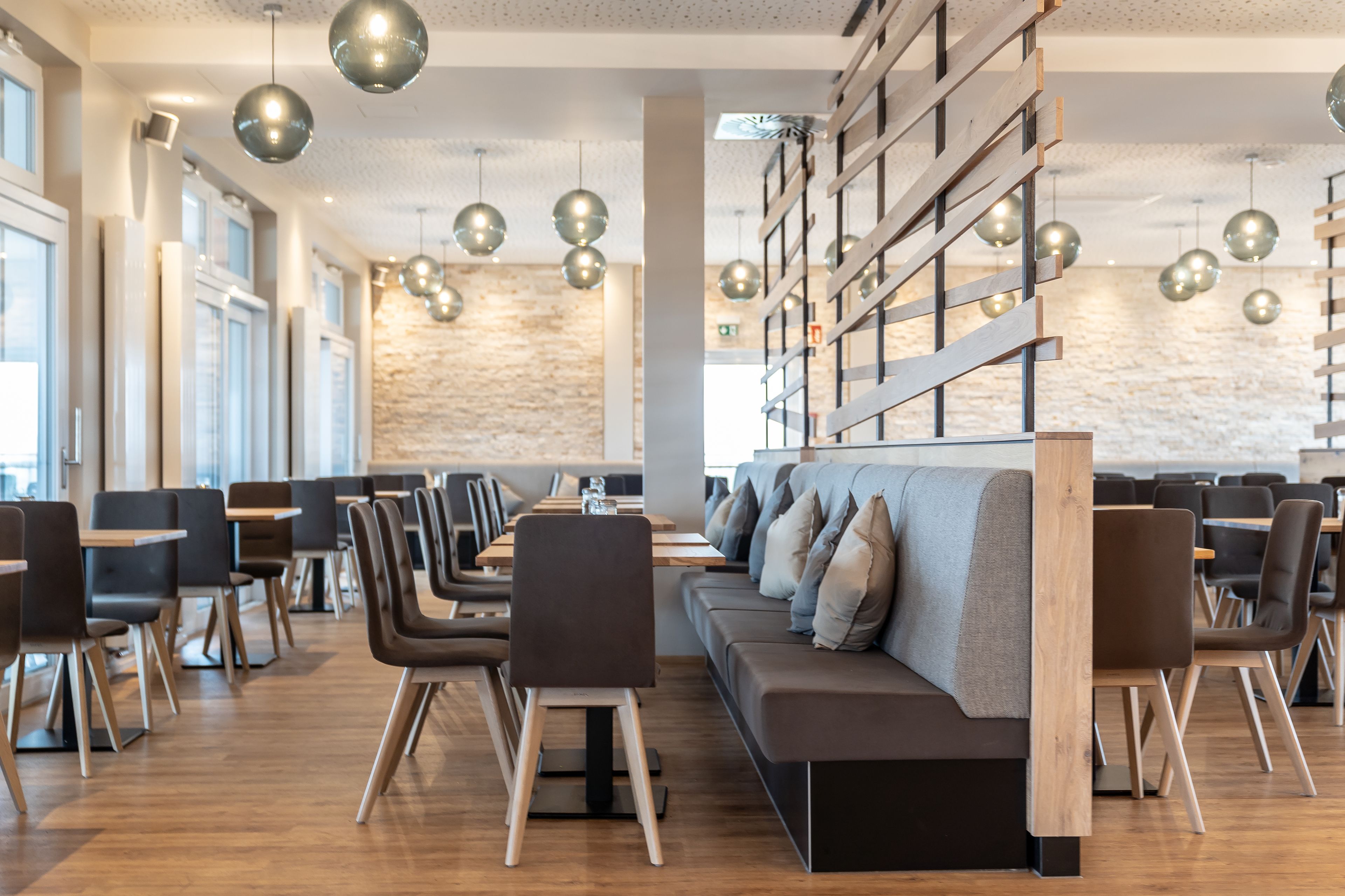 A modern restaurant with wooden tables, black chairs, and a long wooden bench. There are hanging spherical lights and a stone wall in the background.