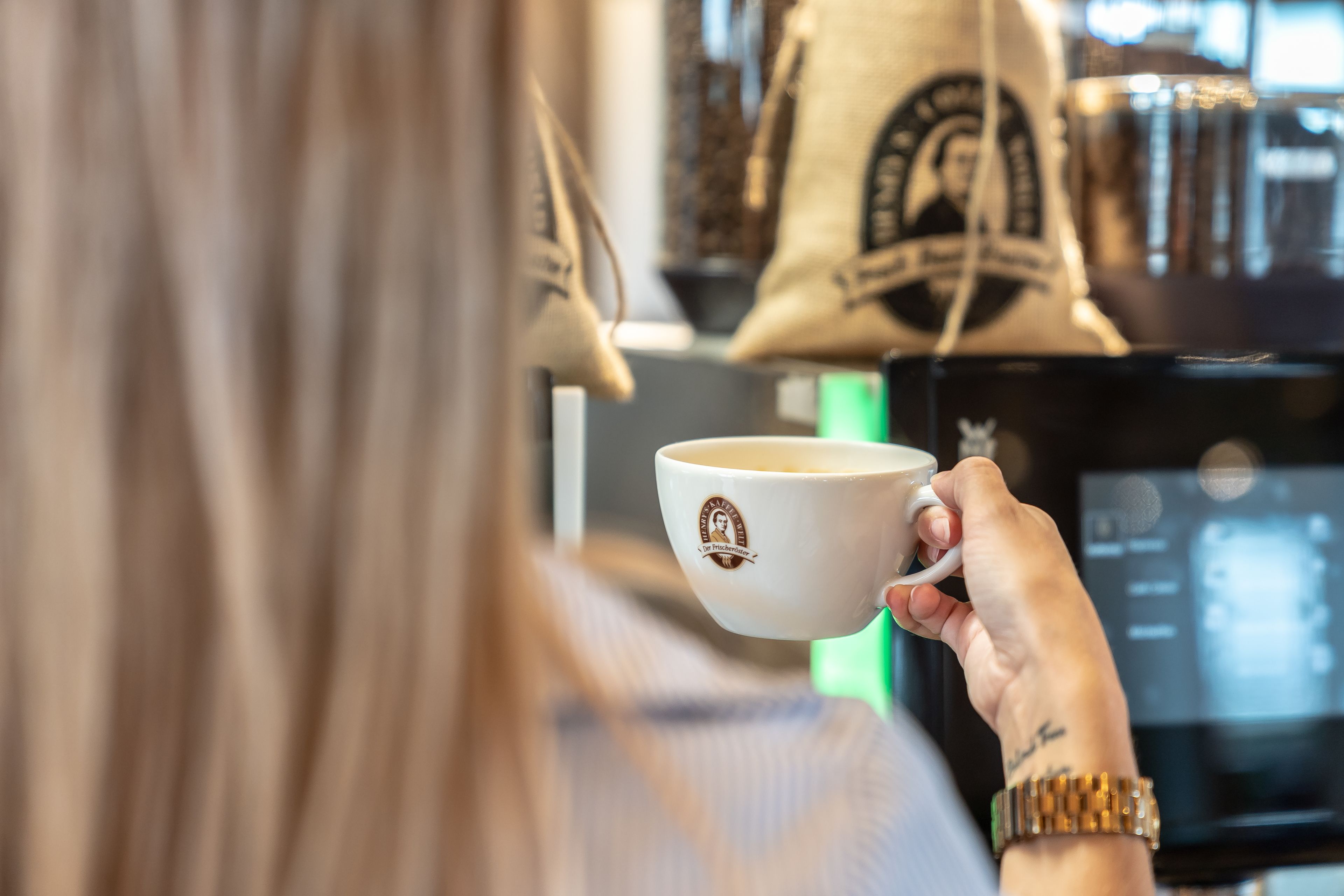 Une personne tenant une tasse ornée d’un logo, dans un décor où l’on aperçoit des sacs suspendus et une machine à café.