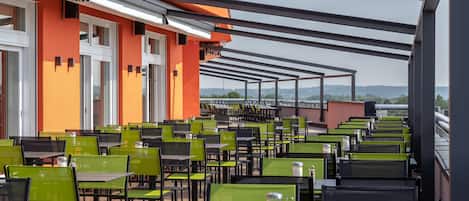 A rooftop terrace with rows of green and black chairs and tables, a building with orange and white walls, and a clear view of the landscape.