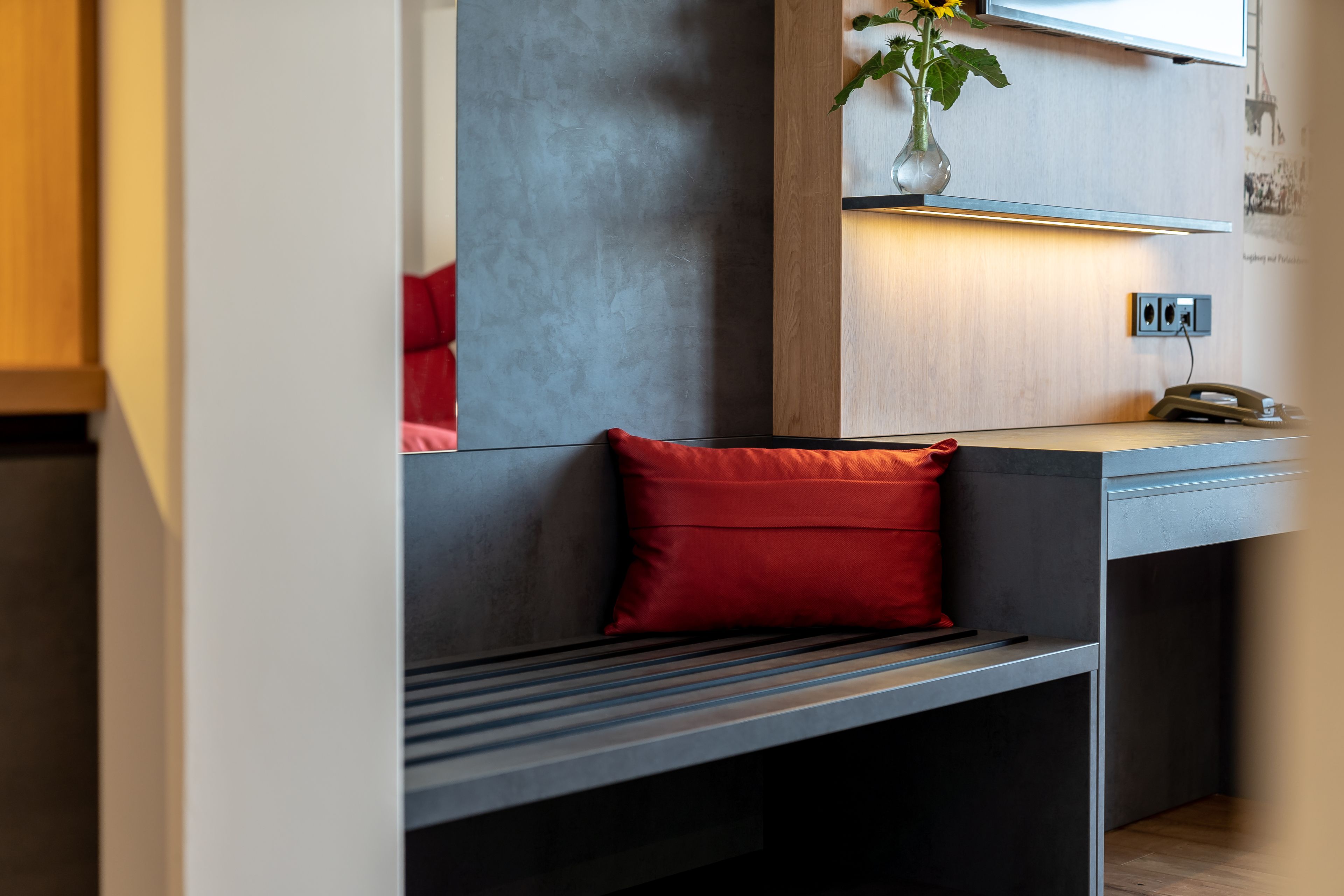 Una habitación moderna con un banco gris, un cojín rojo, un florero con flores y una estantería de madera con un teléfono y una lámpara de pared.