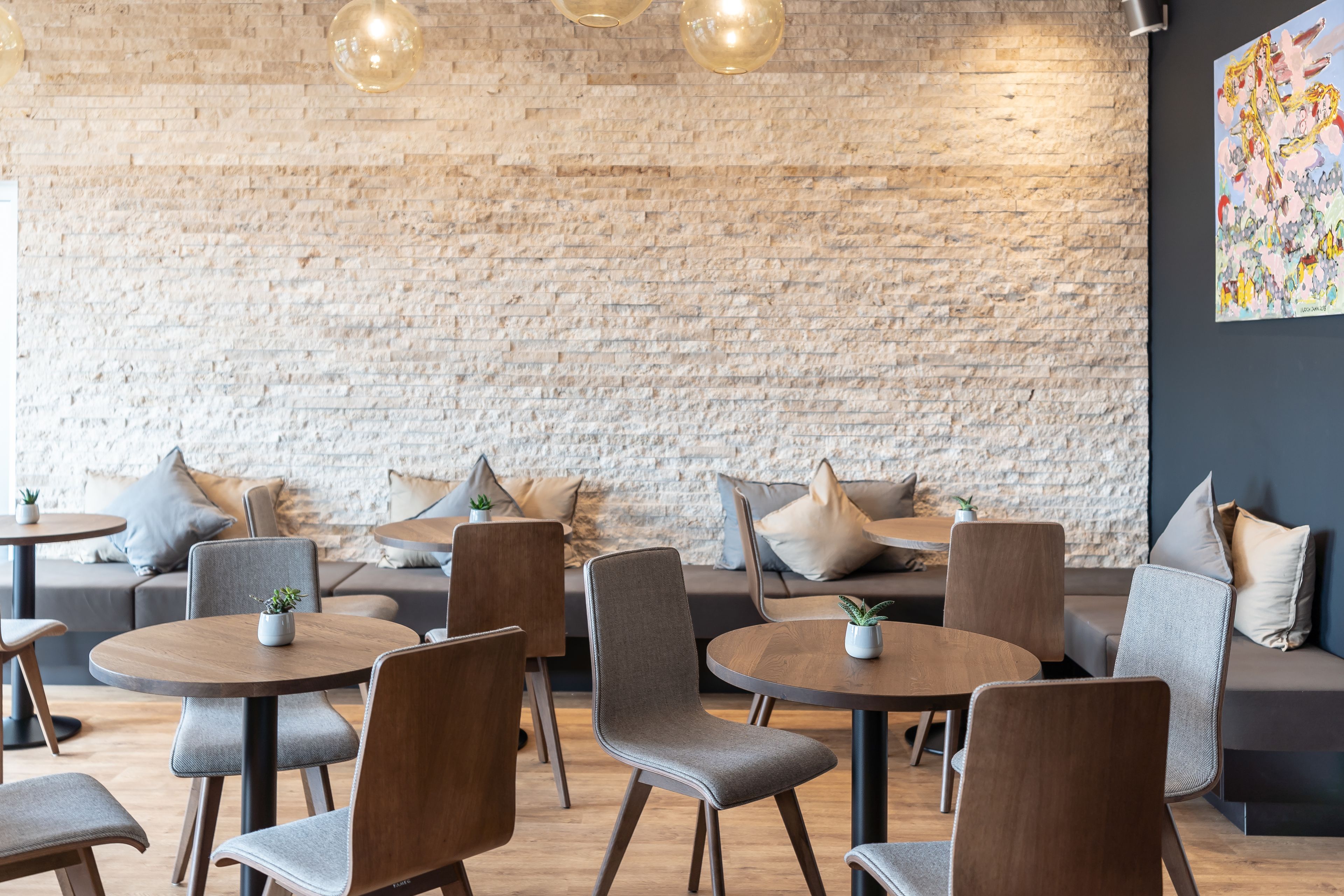 A modern cafe with round tables and grey upholstered chairs, a brick wall, and a colorful abstract painting on the wall.