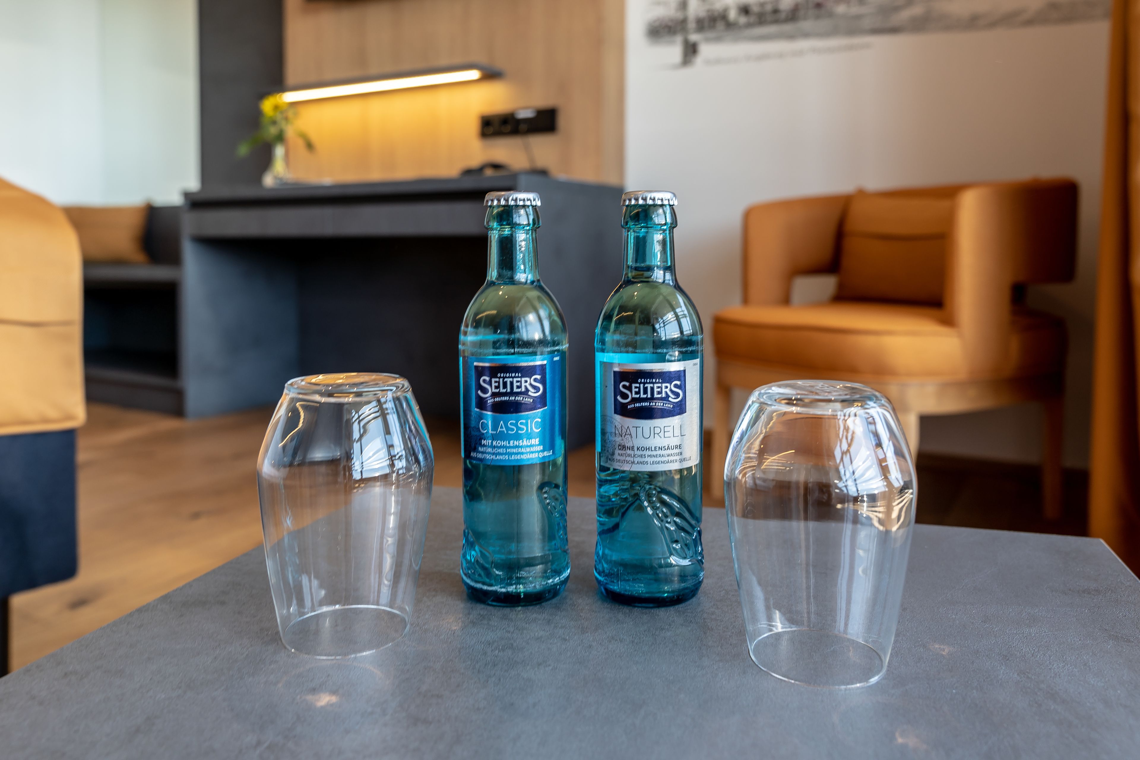 Two bottles of Selters Classic and Selters Naturell water on a table with three empty glasses.