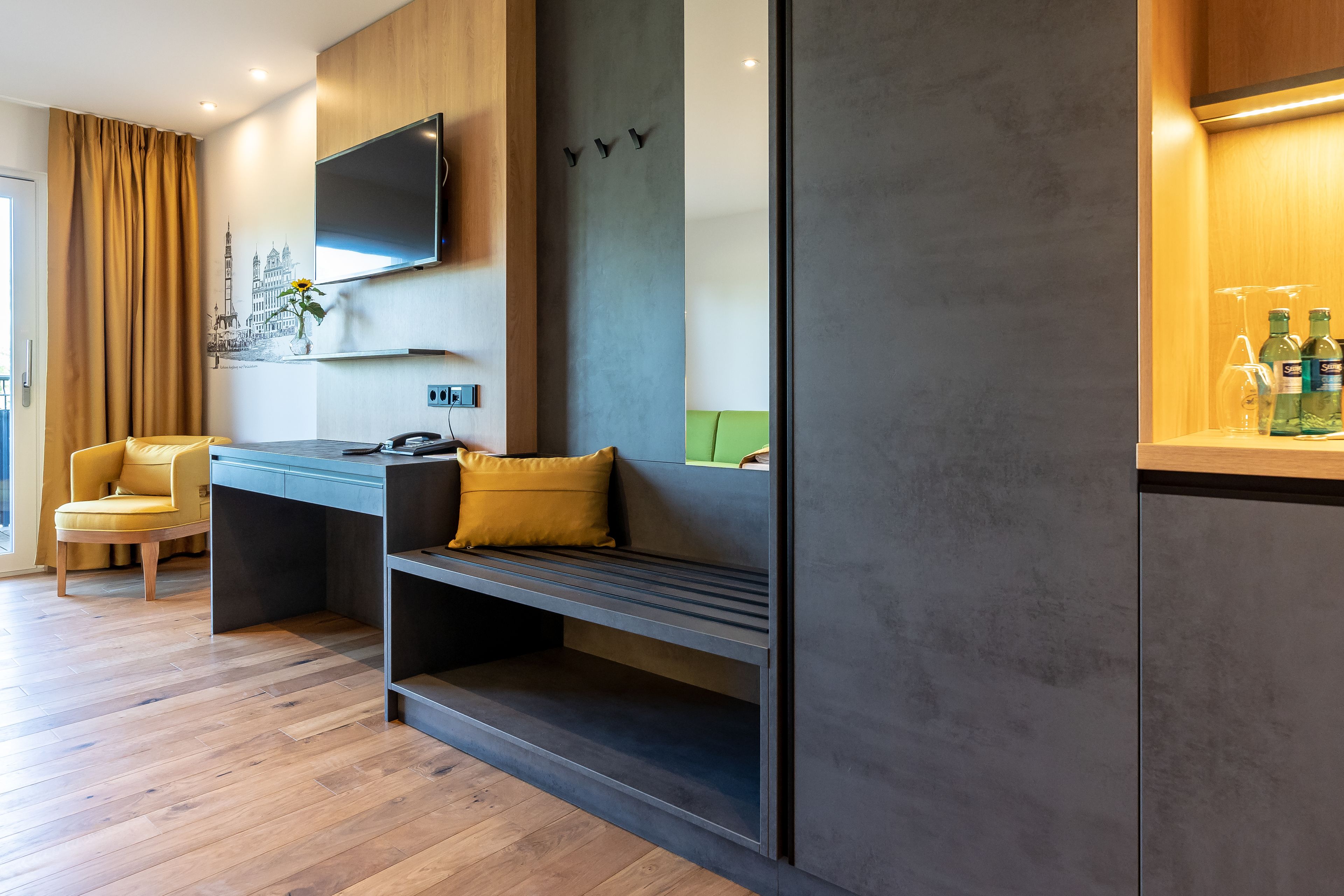 A modern hotel room with a wooden floor, a sofa, a desk with a chair, a flat-screen TV mounted on the wall, and a green accent wall.